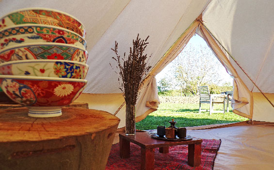 Photo de l'entrée d'une tente vue de puis l'intérieur, avec du mobilier en bois brut et des fleurs
