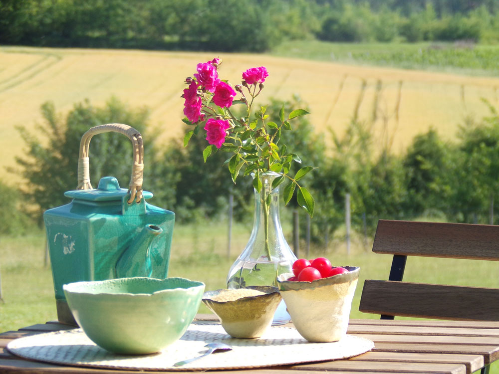 Photo d'un service à thé en céramique avec une branche de rosier dans un vase et les champs en arrière plan